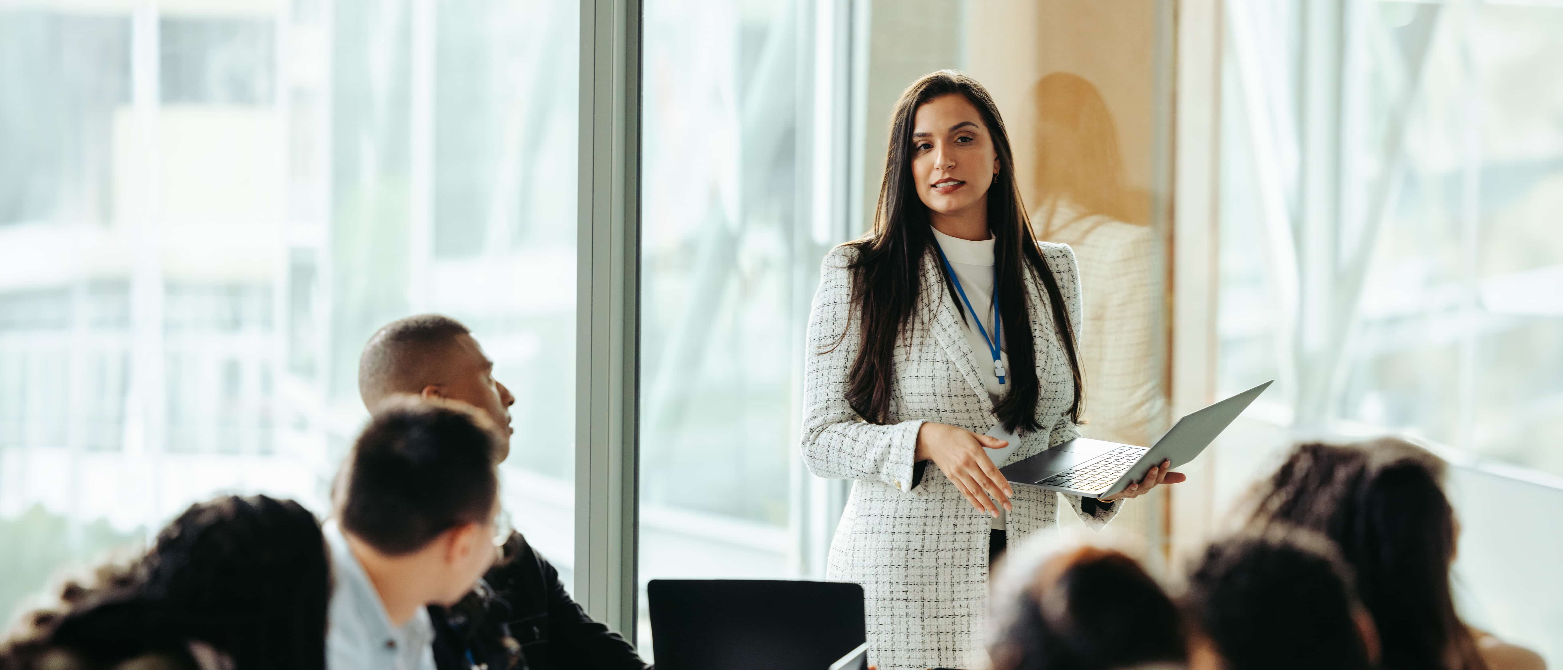 A woman in white business attire speaks to a glass-walled conference room full of people.