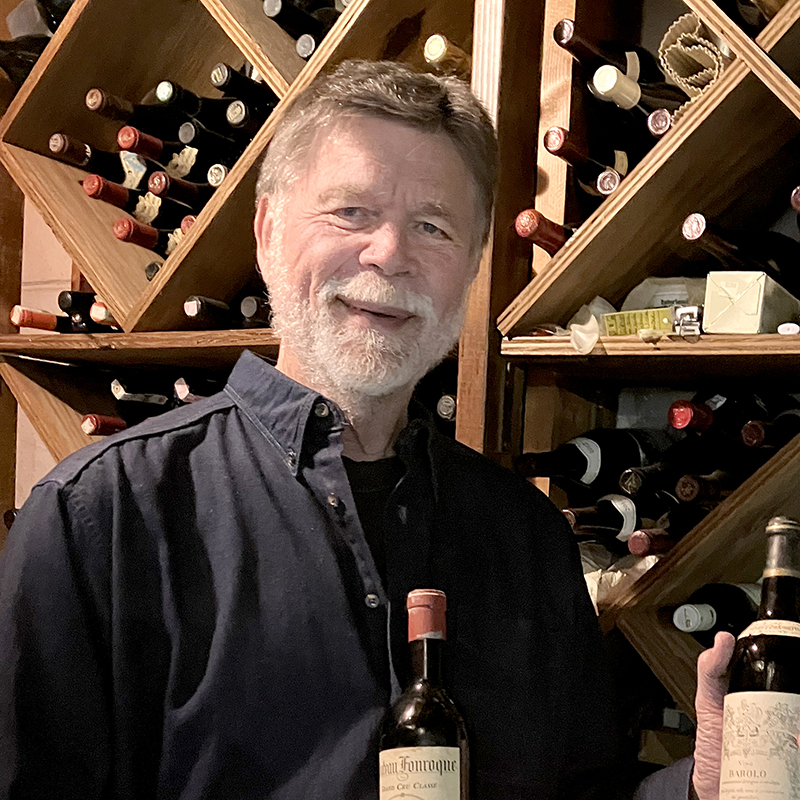 Joe Van Matre, Ph.D., standing in front of his wine cellar holding two bottle of wine.