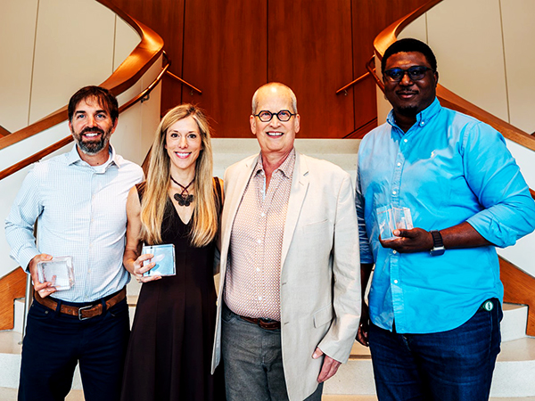 Four business faculty members holding awards in front of stairs