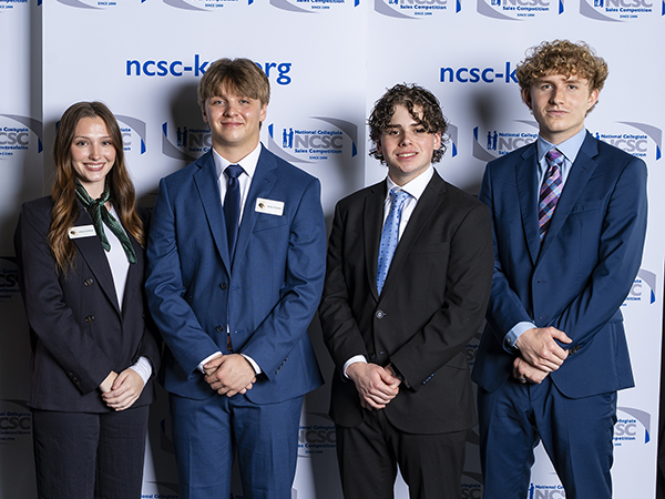 Four smiling students wearing blue suits stand in front of a white backdrop with logos