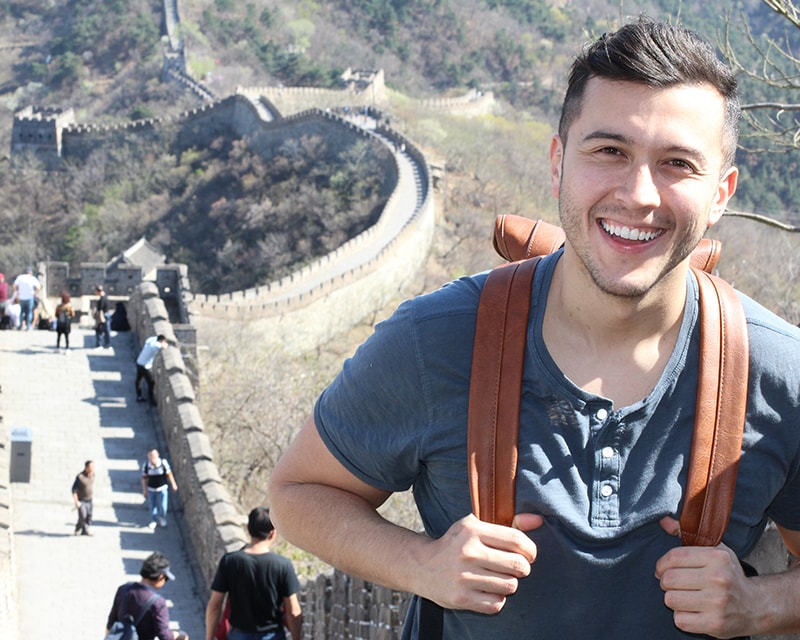 White male student with a backpack smiling atop a segment of the Great Wall. 