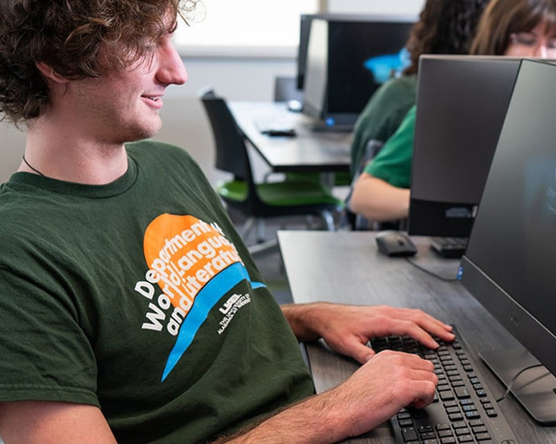 White male student with curly brown hair wearing a department tshirt works at a computer. 