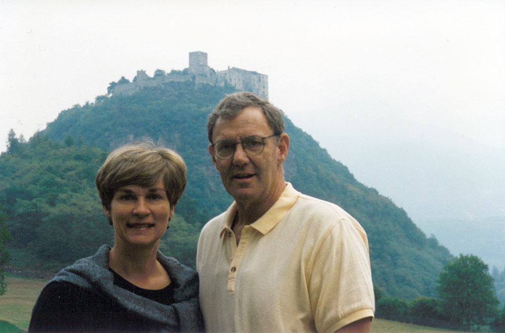 Karen and Lee Barton Chapman standing in front of a wooded hill with a castle on its peak. 