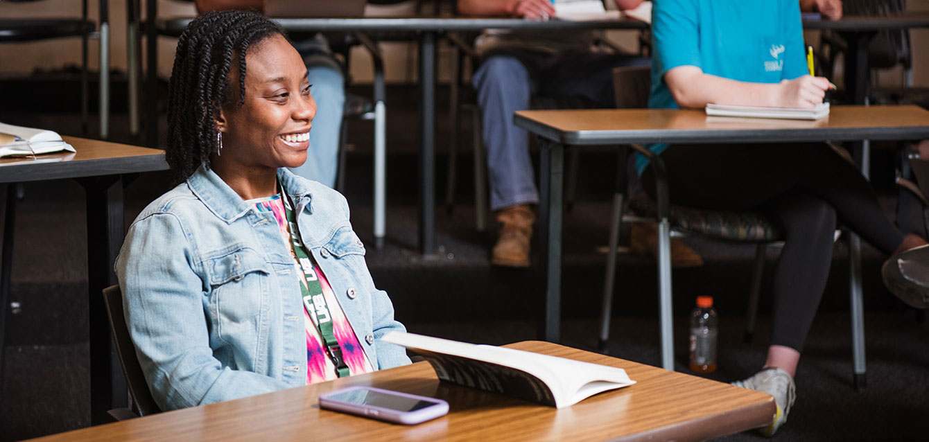 A student sits at a table in her UAB classroom, an open book and her cell phone in front of her. 