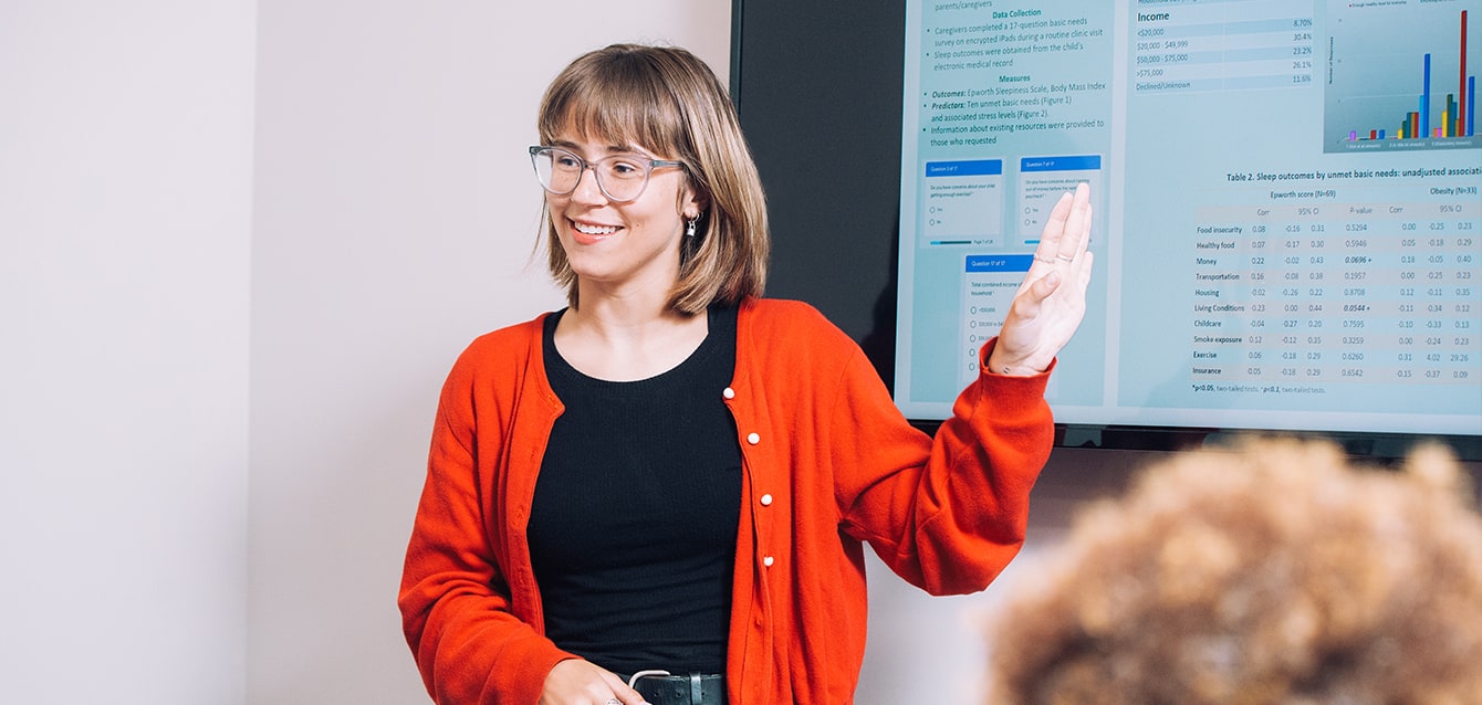 Young woman with shoulder-length straight blond hair, glasses, and a red sweater presents statistical information. 