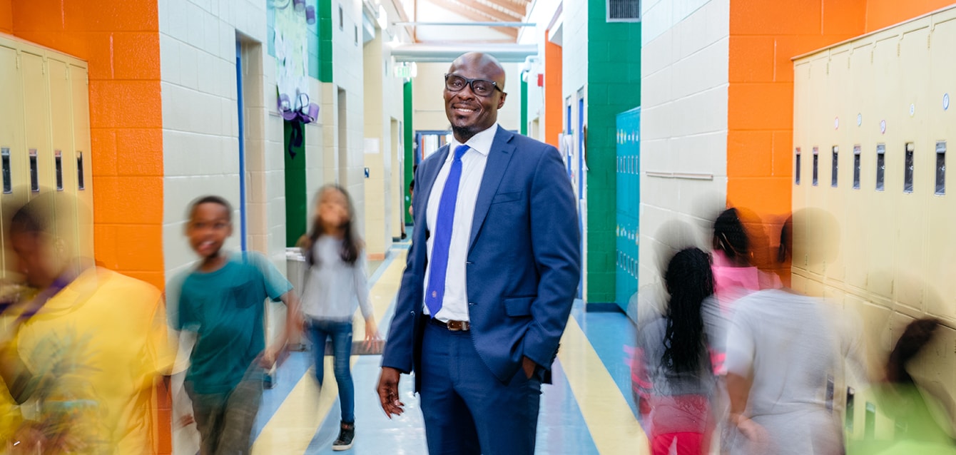 A Black mane in a blue suit and royal blue tie stands in a colorful middle-school hallway, blurred students moving around him. 