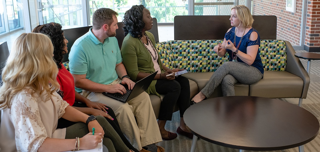 A mix of male and female students sit on a couch listening to a woman speak, taking notes with notebooks, tablets, and laptops.