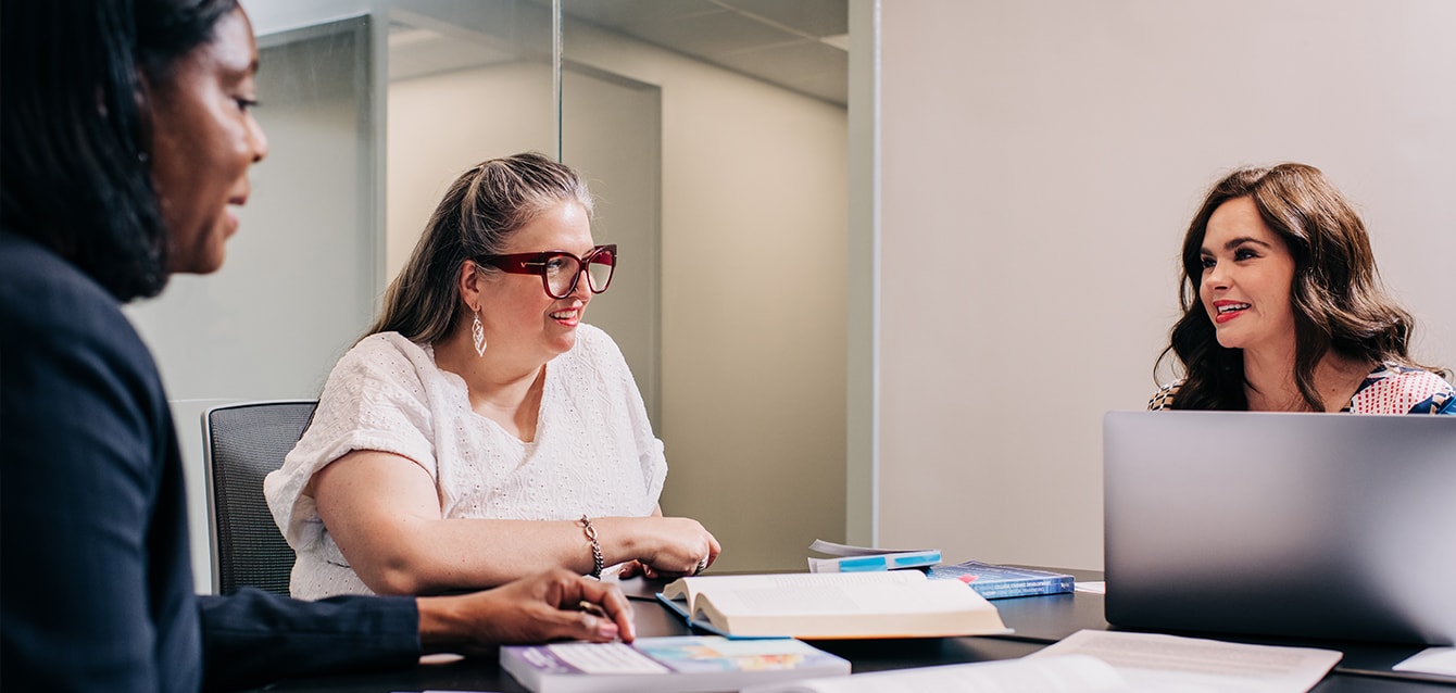 Three women sit around a table in an office-like setting, a laptop on the table in front of one of them. 