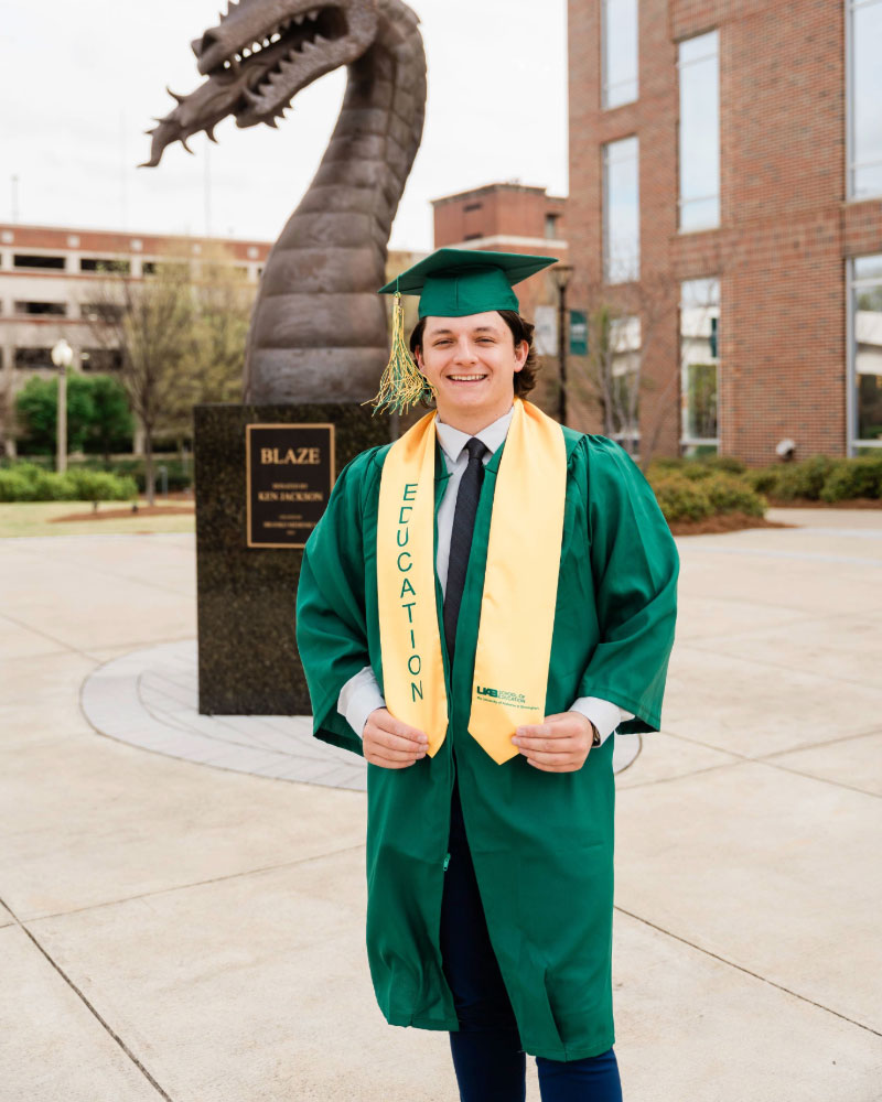 Brandon stands in front of a bronze Blaze statue in his graduation robe and cap. 