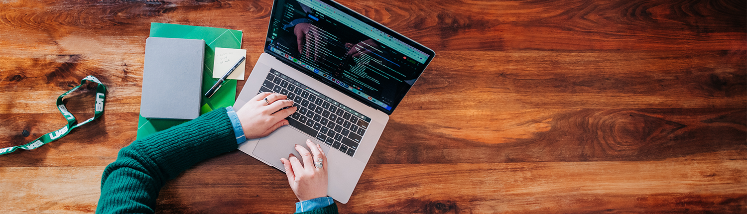 Overhead shot of a pair of female hands working on a laptop sat on a wooden table, surrounded by a green notebook, post-its, pen and a UAB lanyard
