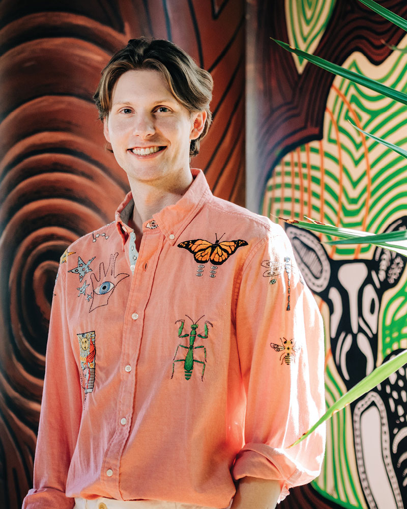 A portrait of medical student Matthew Kiszla. He wears a salmon shirt with hand-embroidery and is pictured against a brightly colored mural background. 