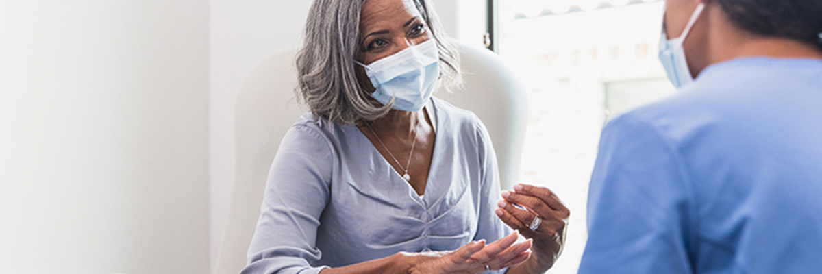 A senior woman, wearing a protective face mask, talks with a female nurse during a medical appointment.