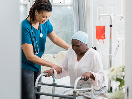 Young female home healthcare professional helps an elderly woman get out of bed by using a walker. The woman is wearing a cap on her head. An iv drip is in the background.