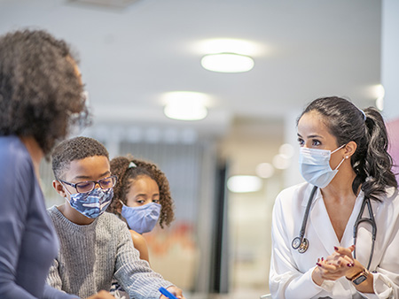 A doctor interacts with a mother and her two children in a facility. They are all wearing masks.