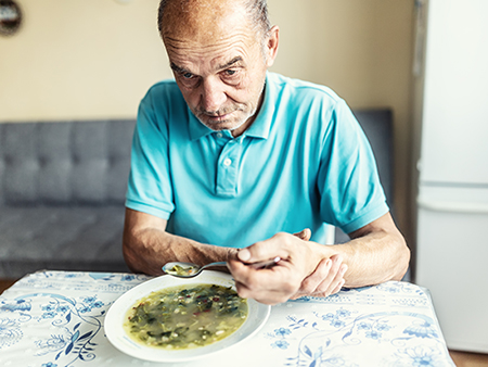 Senior man with Parkinsons disease holds his arm with a hand, trying to eat a soup.