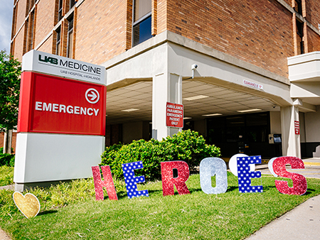 Exterior of UAB Hospital-Highlands showing the Emergency entrance with decorative lettering that reads "Heroes" staked in the grass in patriotic colors during the COVID-19 (Coronavirus Disease) pandemic; blue sky and white clouds are above, April 2020.