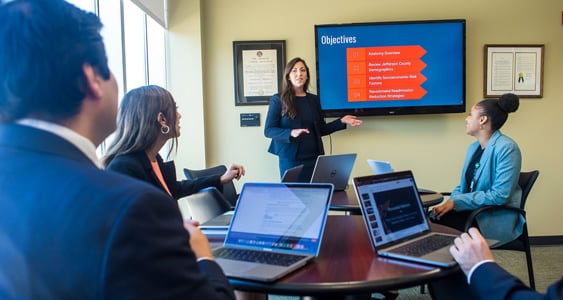 Woman standing in front of powerpoint aand presenting to a agroup of people.